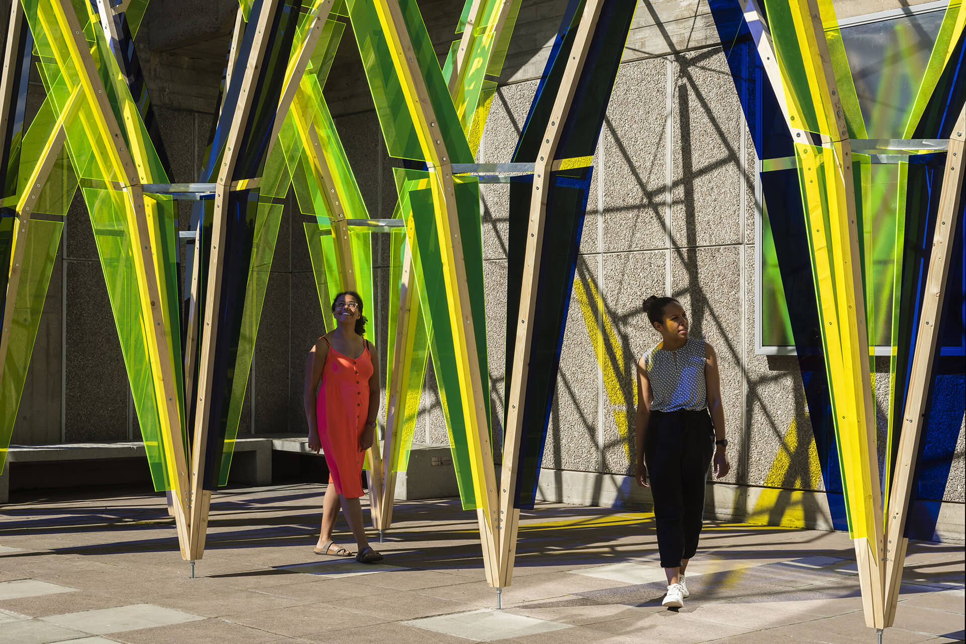 Jyll Bradley's The Hop sculpture, installed outside The Hayward Gallery at Southbank Centre. It is made of tall beams of yellow and blue perspex reaching up towards the sky, at approximately 12 feet tall. Two women are positioned between the beams, looking at the sculpture.