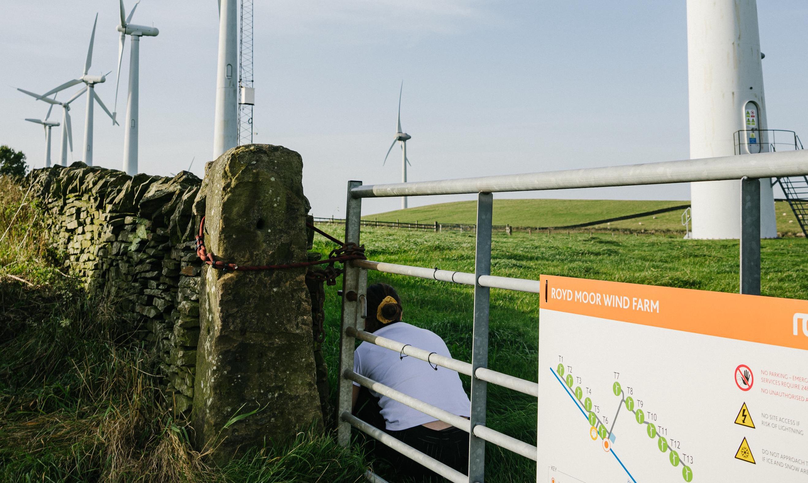 Composer Sara Rahman crouches just inside the gate of a wind farm in Yorkshire. The wind turbines are visible in the image and span a lush green pasture bordered by a jagged stone wall.